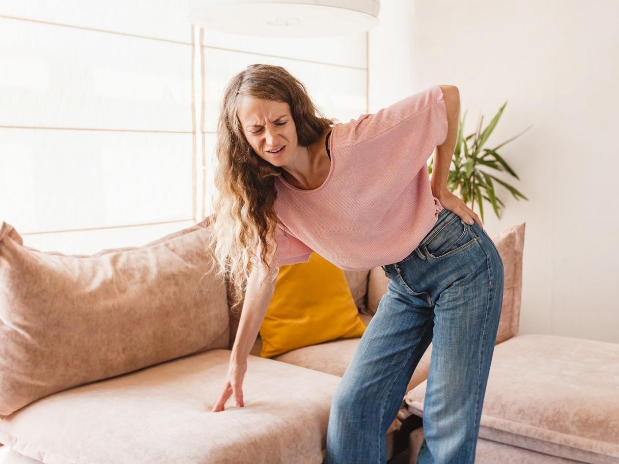 Chronic back pain. Adult woman is holding her lower back, while standing and suffering from unbearab